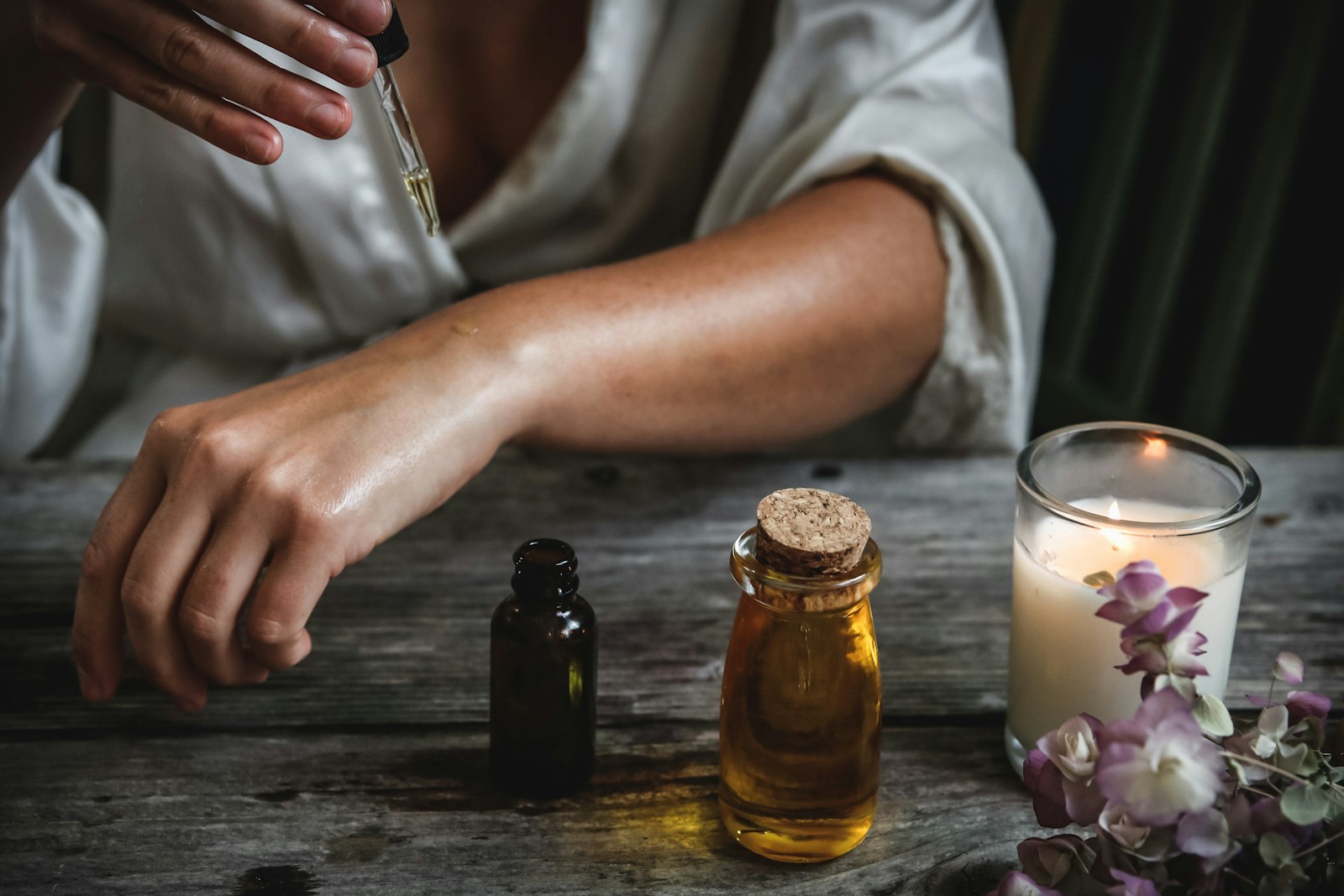 A person applying oil from a glass dropper onto their arm at a table